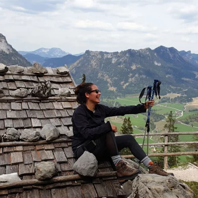 Photo of a woman in sunglasses, smiling, sitting on the roof of a cabin, holding walking sticks. On the background you see the alps.