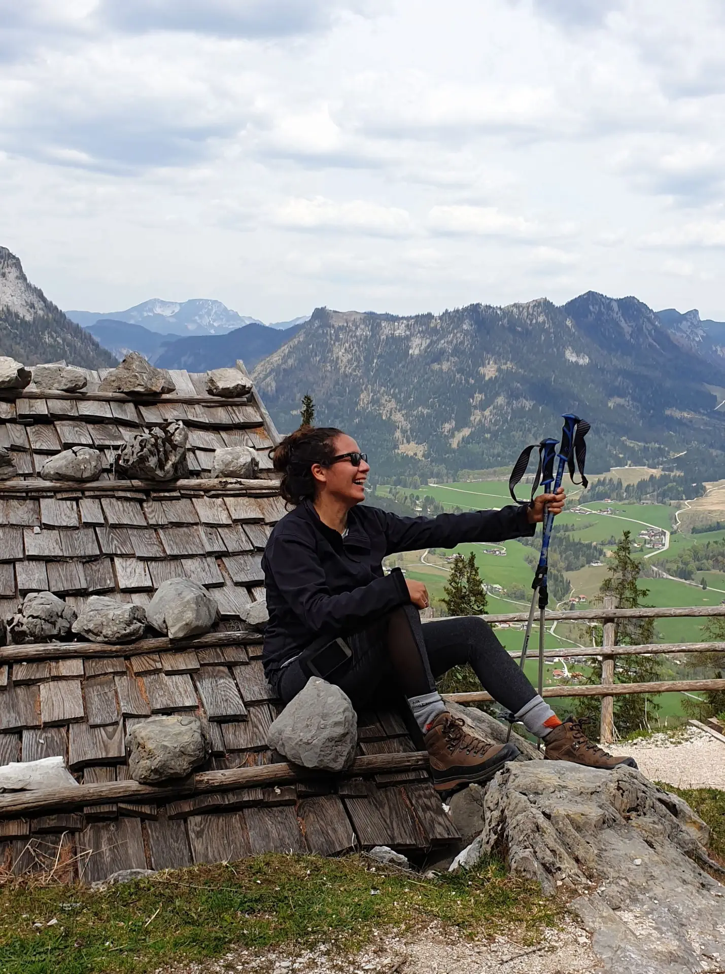 Photo of a woman in sunglasses, smiling, sitting on the roof of a cabin, holding walking sticks. On the background you see the alps.